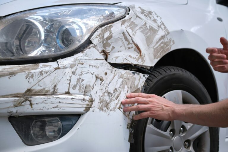 Driver hand examining dented car with damaged fender parked on city street side. Road safety and